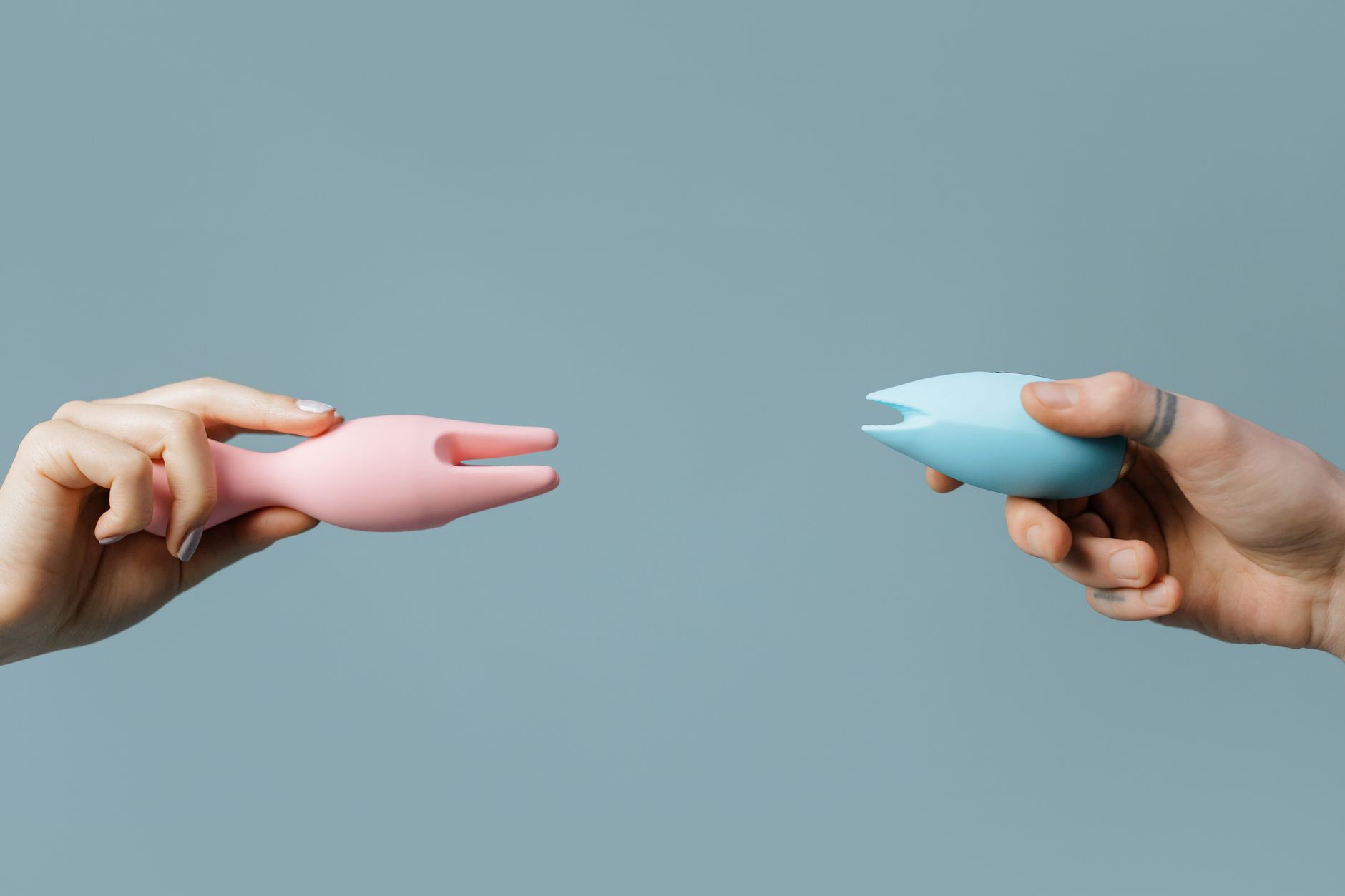 Duas mãos segurando vibrador silicone rosa e azul contra fundo pastel claro