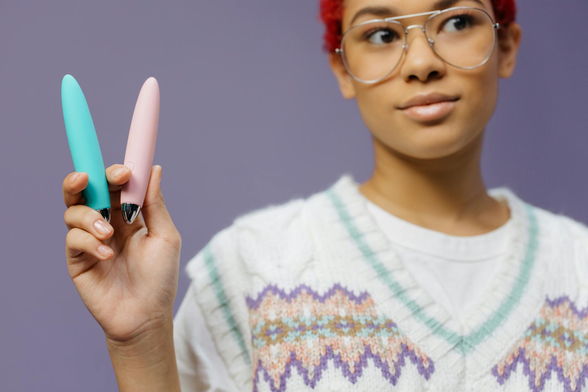 Mulher jovem com óculos segurando dois vibrador coloridos contra um fundo roxo